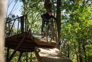 A female mountain biker is riding down a steep bridge onto a big flat rock.